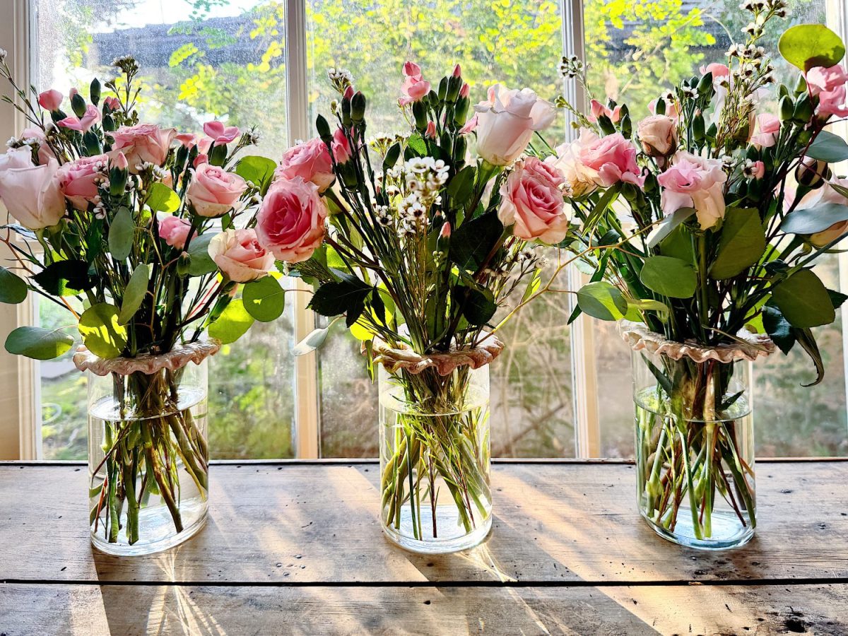 Three glass vases filled with pink roses and greenery on a wooden table. Sunlight streams through a window in the background.