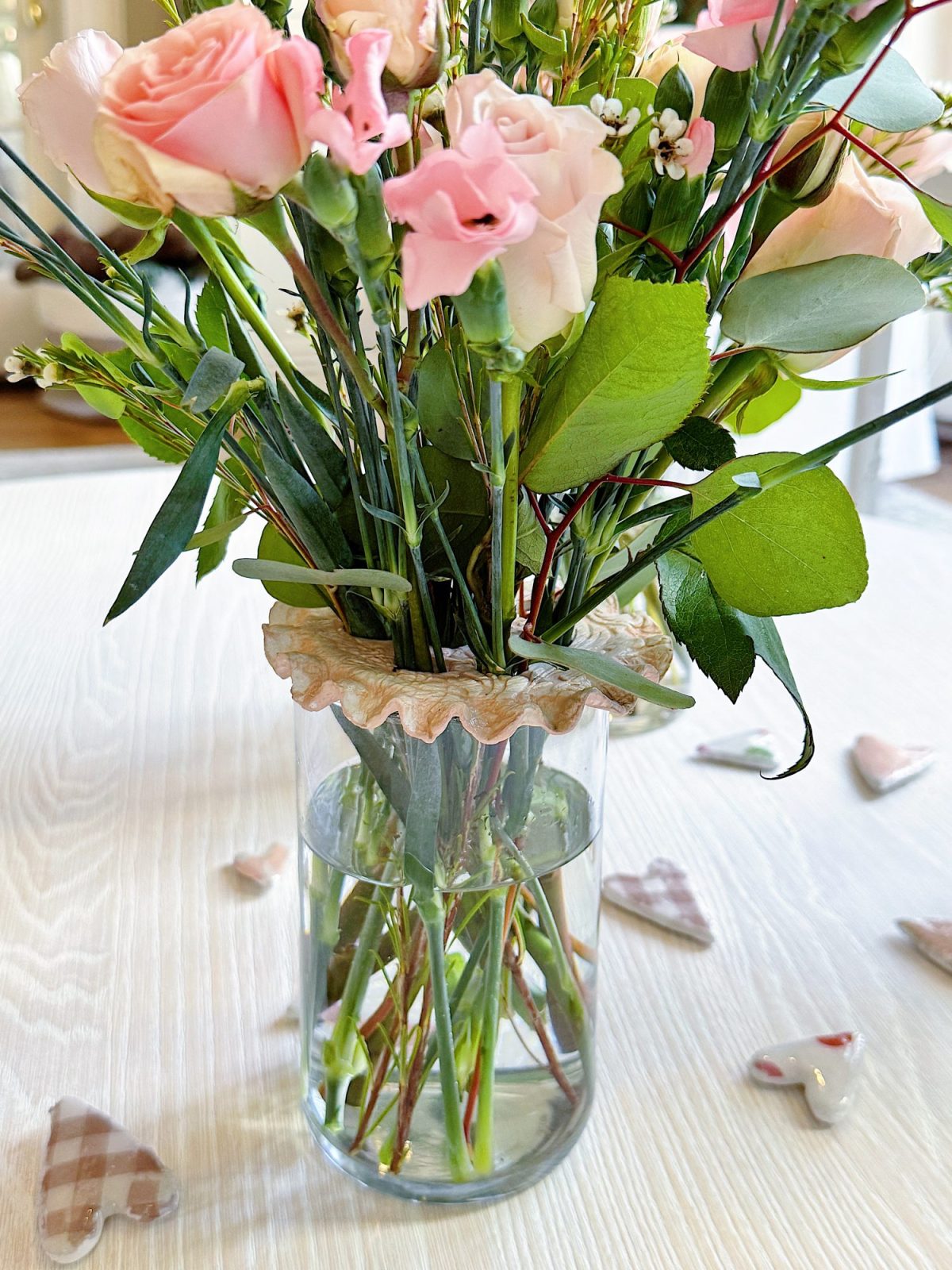 A glass vase with pink roses, flower frogs, and green foliage on a white surface, surrounded by small heart-shaped decorations.