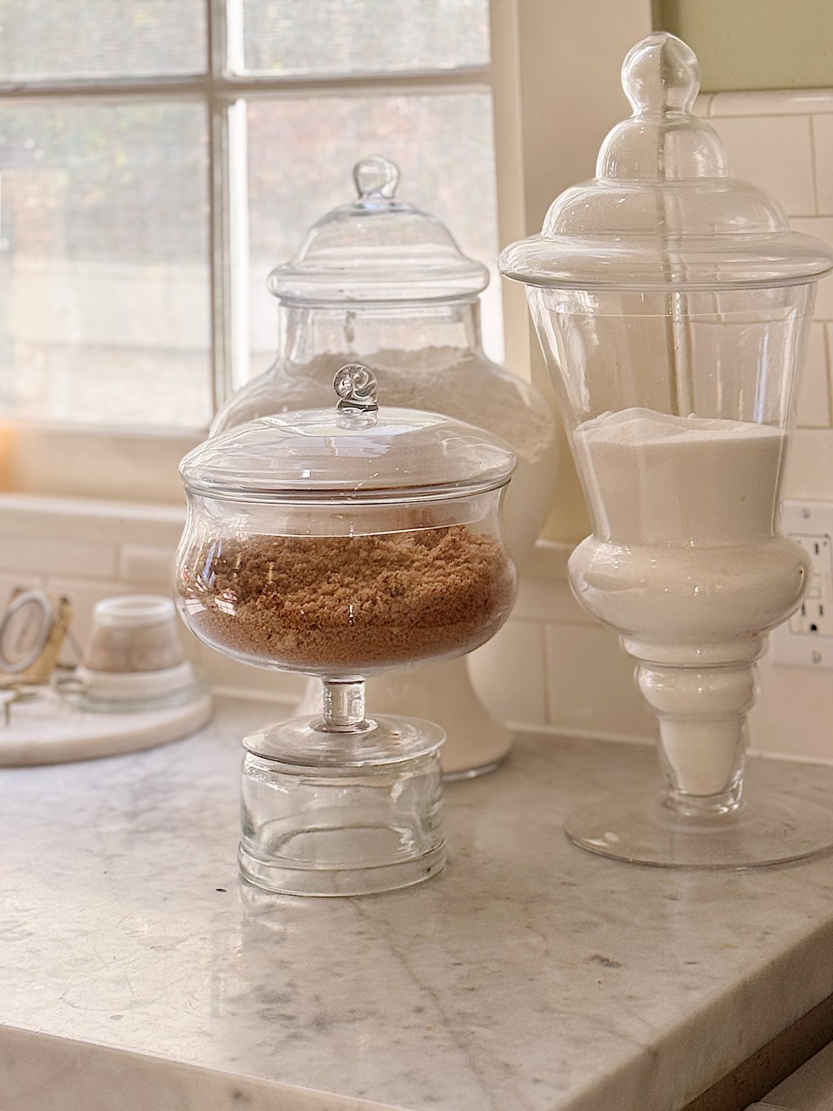 Glass jars filled with brown sugar, flour, and white sugar sit on a marble countertop in front of a window.