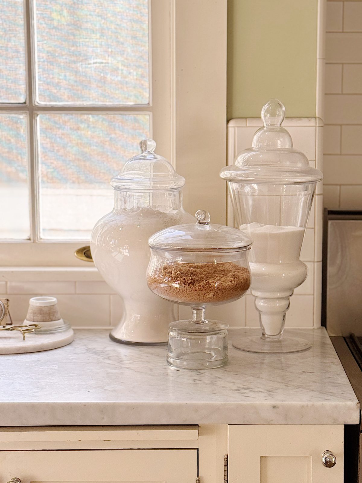Glass jars filled with flour, brown sugar, and white sugar are neatly arranged on a marble kitchen countertop near a window.
