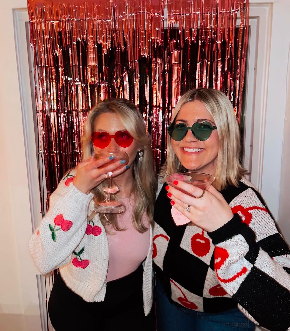 Two women in heart-shaped sunglasses hold drinks in front of a shiny red backdrop. They wear cardigans with heart and cherry designs.