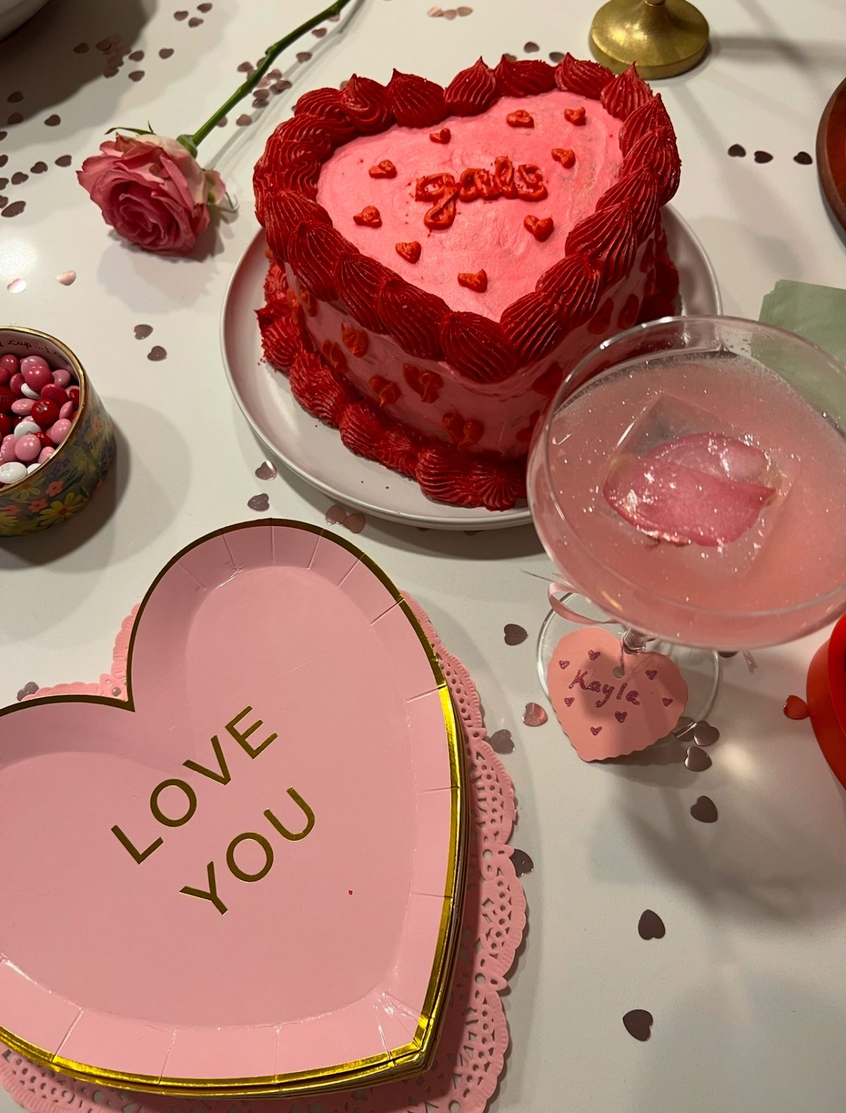Heart-shaped pink cake with red icing and "Jolie" written on top, next to heart-themed decor including a pink drink, a "LOVE YOU" plate, and a single pink rose.