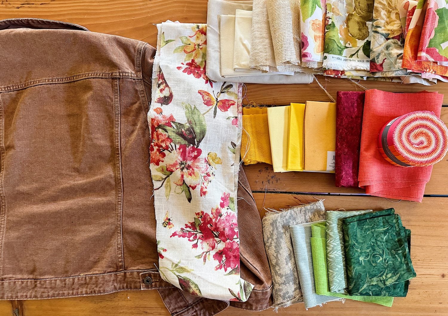 A brown jacket next to assorted fabric swatches in floral, solid, and patterned designs, arranged on a wooden table.