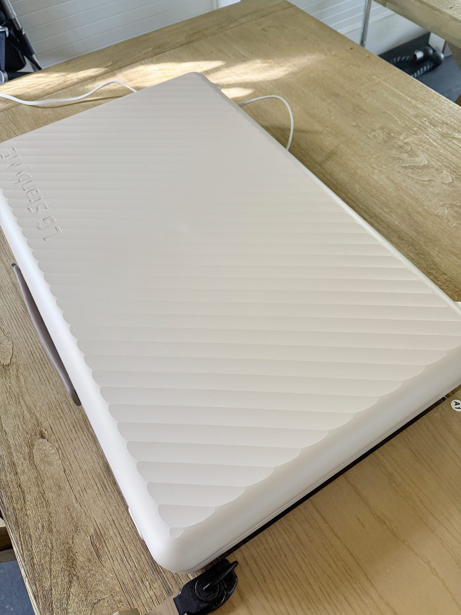 A white suitcase with a ribbed texture is resting on a wooden surface. A small portion of a black wheel and handle are visible at the edge.