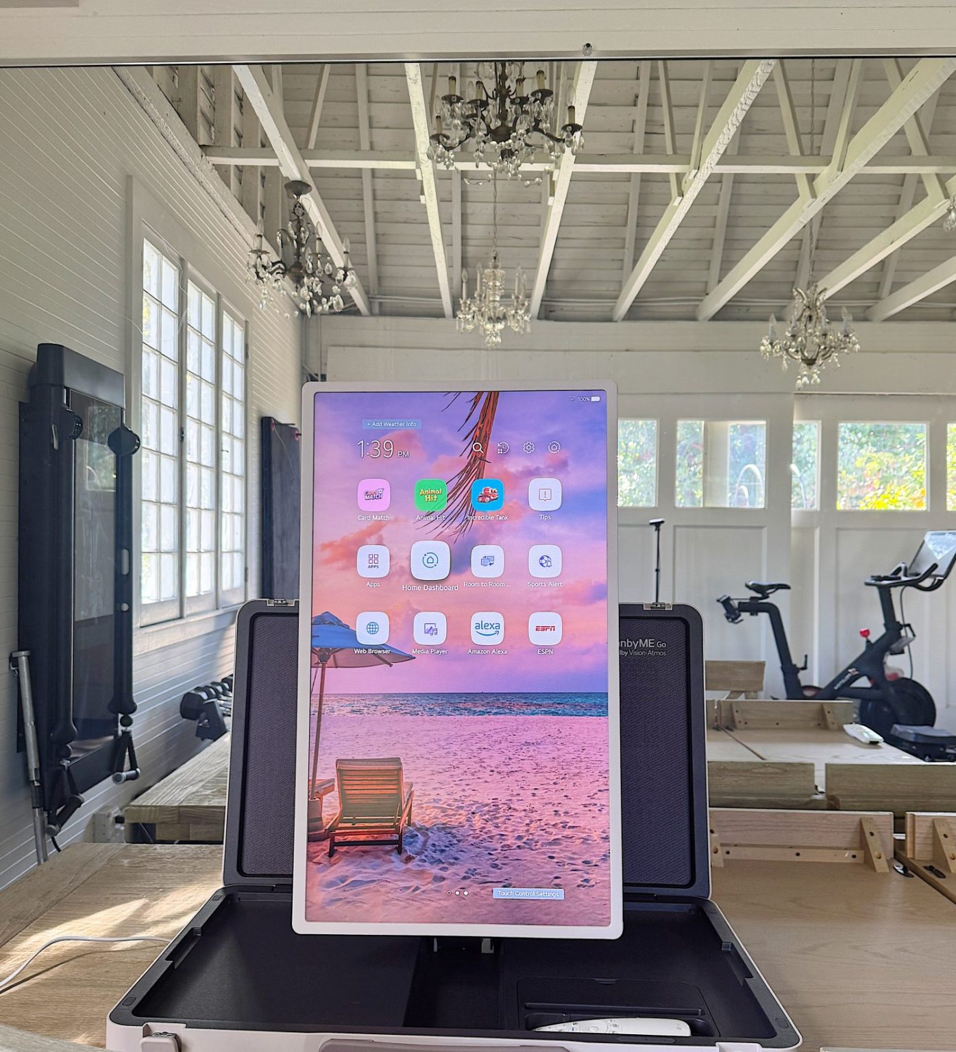 A digital display shows a beach-themed interface with app icons, placed in a bright room with white walls, chandeliers, and gym equipment like exercise bikes in the background.