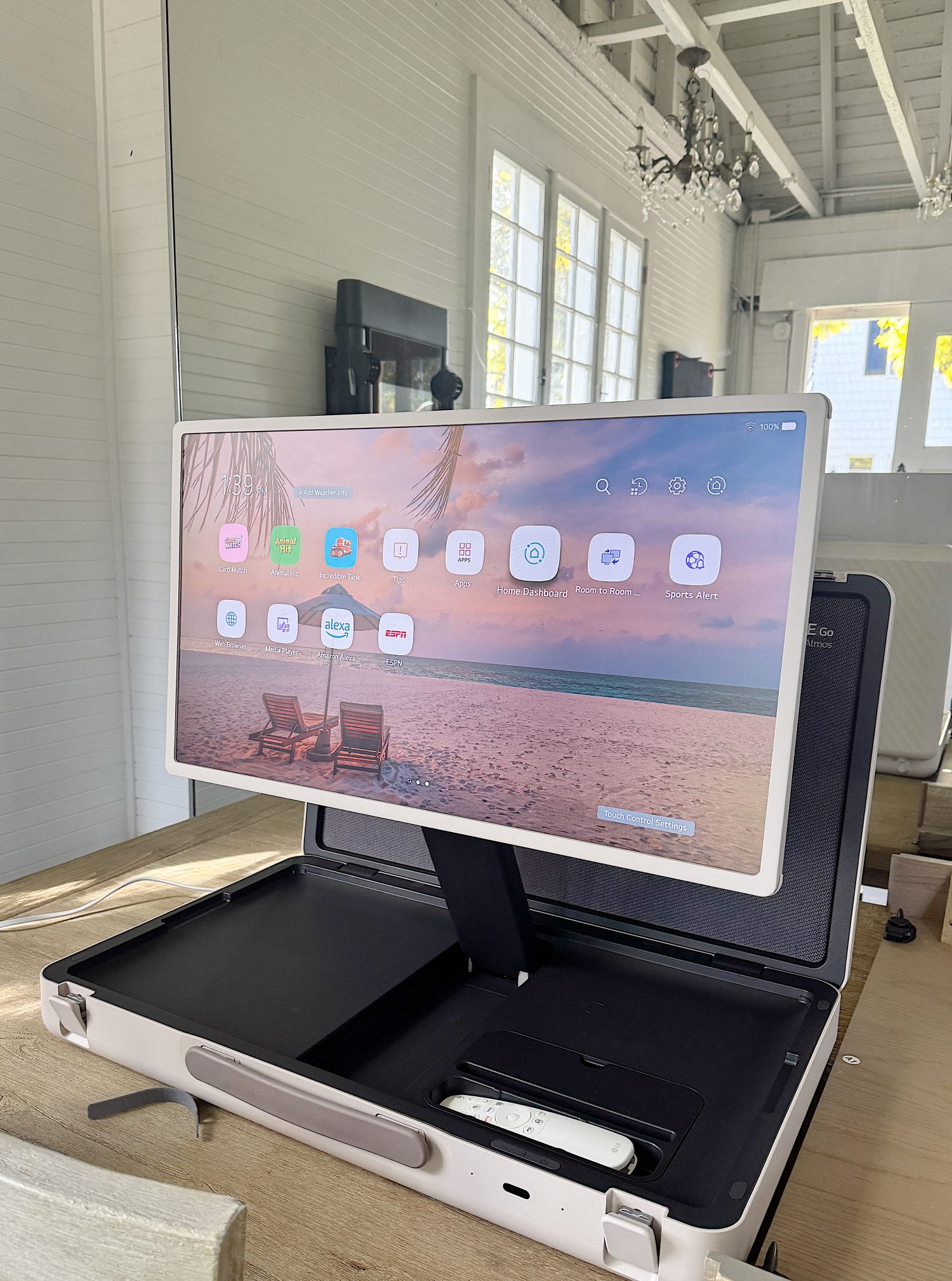 A monitor displaying a palm beach background with apps is attached to an open case on a wooden desk in a bright room with white walls and large windows.