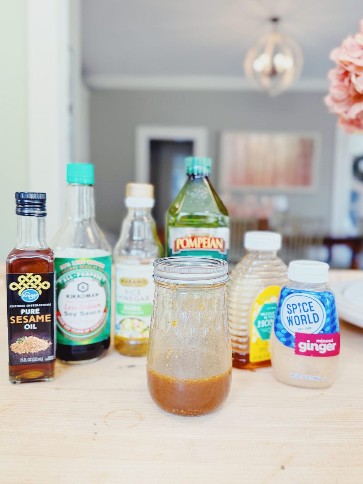 A jar of mixed marinade on a wooden table, surrounded by bottles of sesame oil, soy sauce, vinegar, olive oil, honey, and minced ginger.