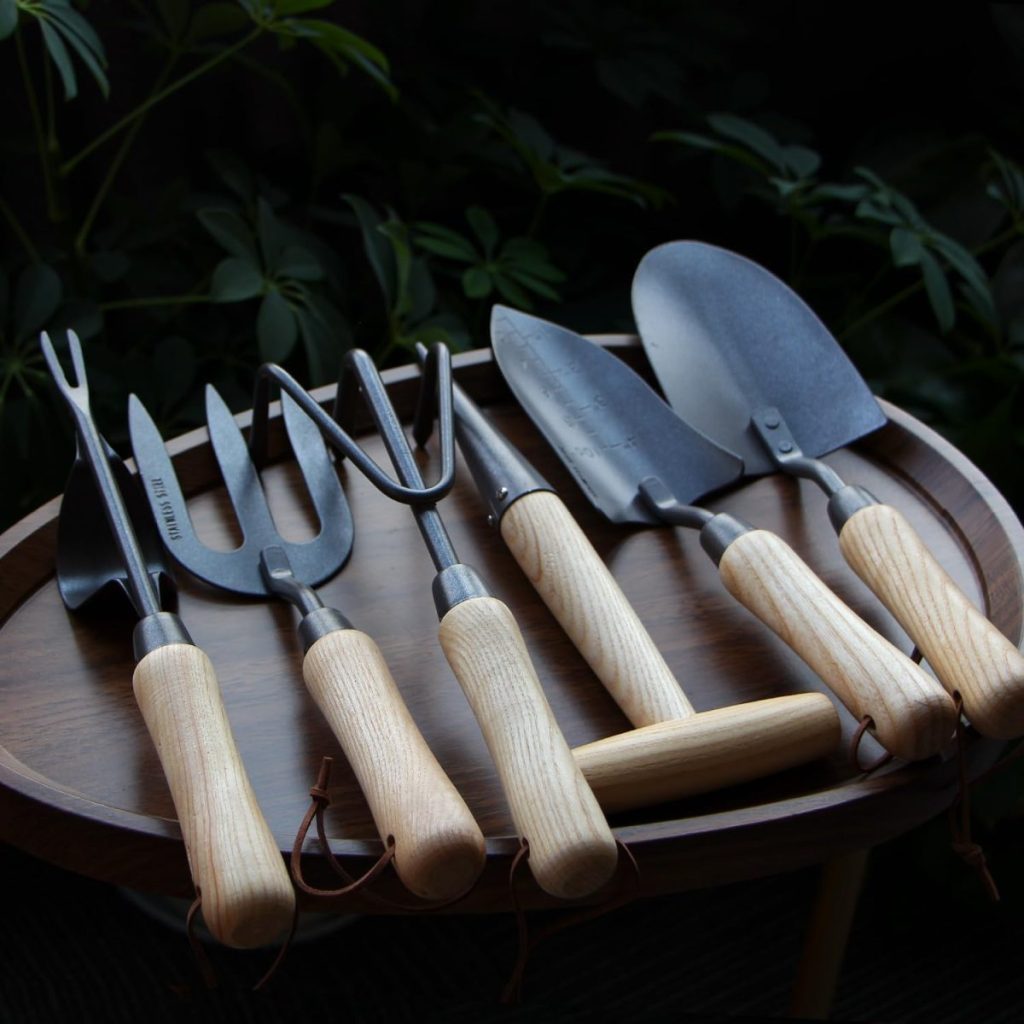A set of six gardening tools with wooden handles and metal heads arranged on a round wooden tray&mdash;perfect for tending to your plants or organizing supplies for your hanging planters.