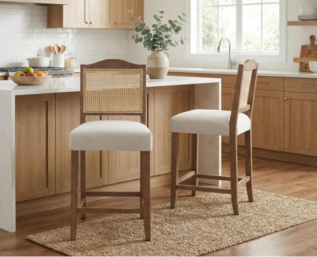 Two wooden bar stools with cane backs and upholstered seats sit by a white kitchen island, complementing the modern kitchen&rsquo;s light wood cabinets and woven rug.