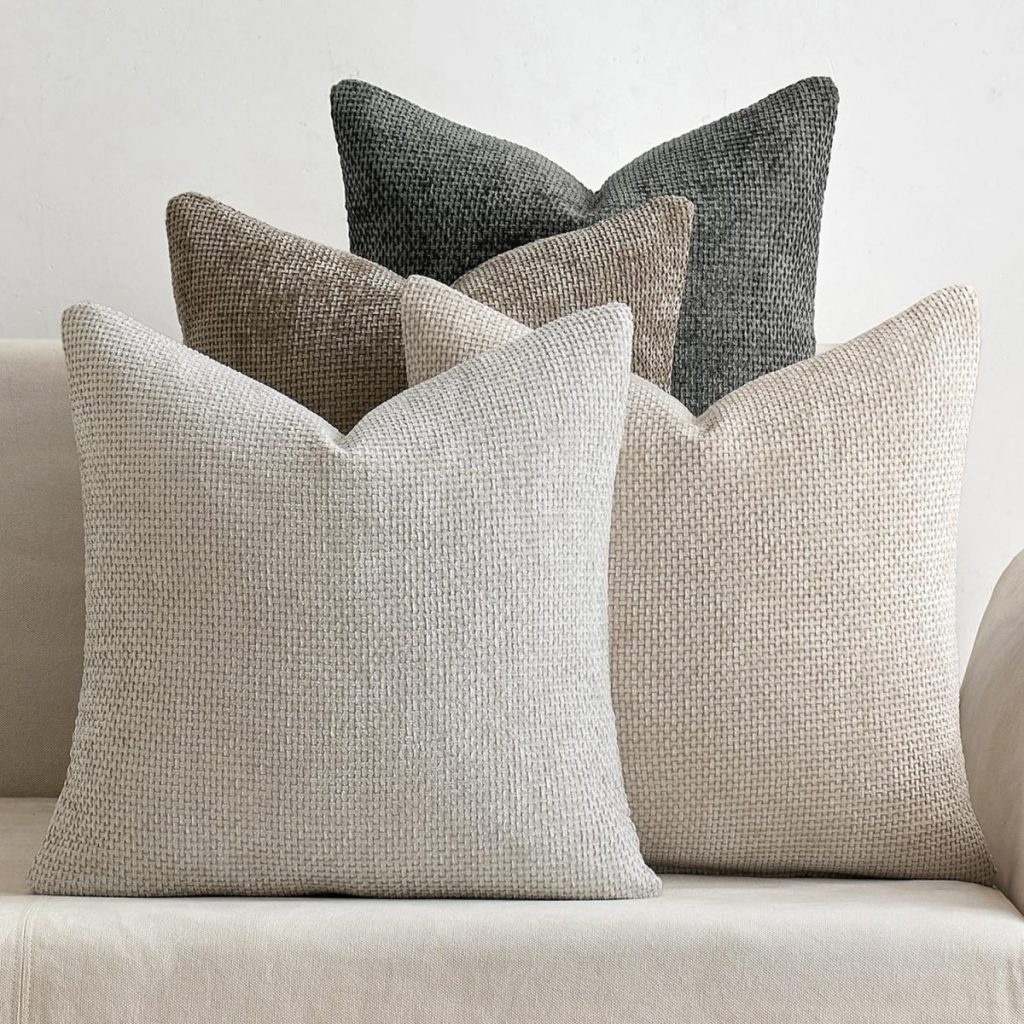 Five textured throw pillows in neutral shades of beige, brown, and gray&mdash;each featuring Pottery Barn pillow covers&mdash;are arranged on a light-colored sofa against a plain background.