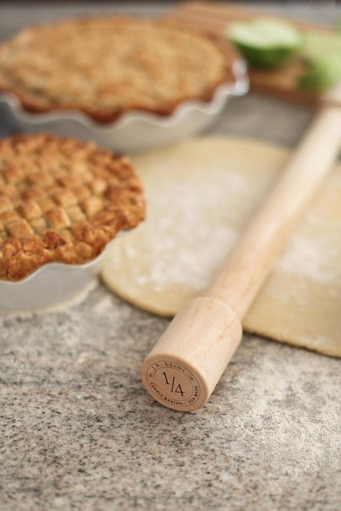 A rolling pin rests on a floured surface next to an unbaked pie crust, with two baked pies in the background, their crusts subtly adorned with Easter Egg designs.