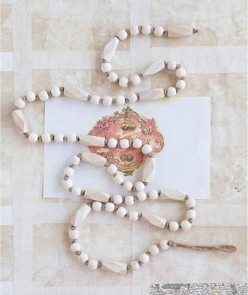 A strand of wooden beads with geometric shapes is coiled on a paper with a pink decorative design, set against a light grid-patterned background—perfect as charming spring decor.