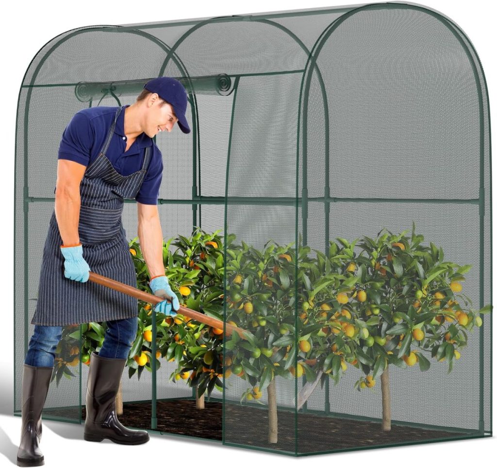 A man wearing boots, gloves, and an apron tends to fruit trees with a hoe inside a green mesh greenhouse, cultivating his kitchen garden.