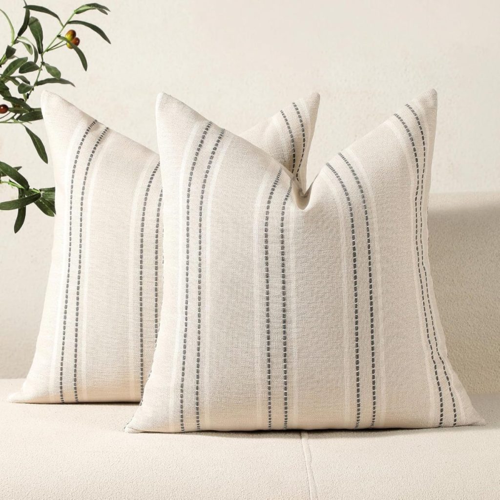Two cream-colored decorative pillows with thin black vertical stripes, featuring Pottery Barn pillow covers, are placed on a light-colored sofa, with a plant partially visible in the background.