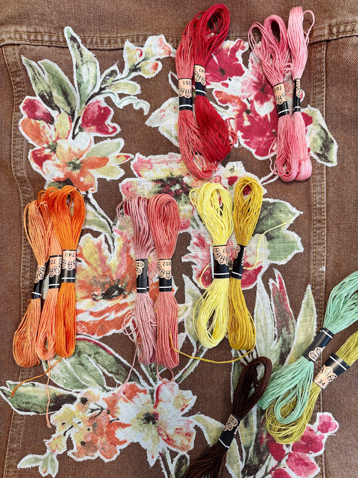 Assorted embroidery threads in various colors, including red, pink, orange, yellow, and green, are arranged on floral-patterned brown fabric.