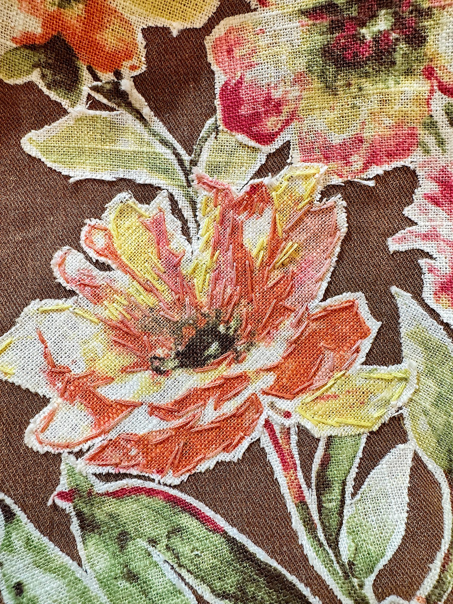 Close-up of a floral pattern on fabric, featuring a large orange and yellow flower with green leaves on a textured brown background.