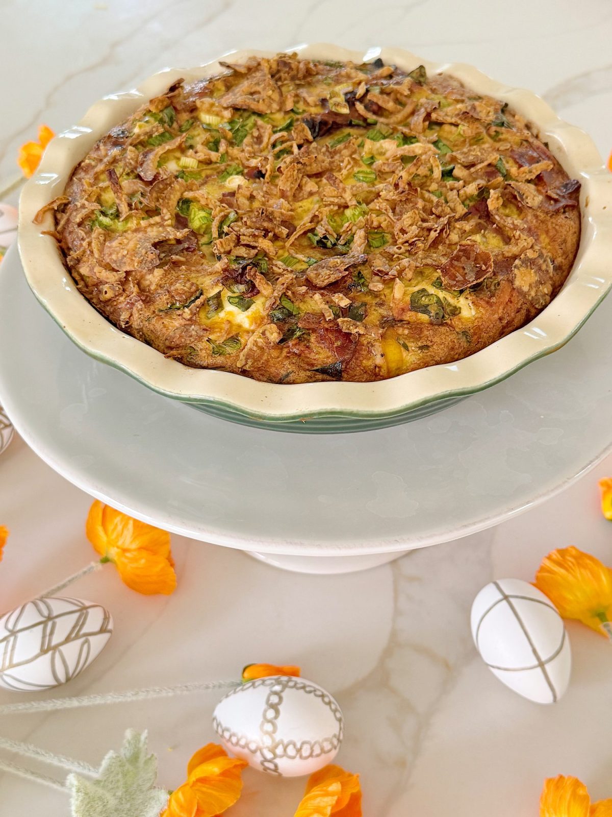 A savory pie on a stand, topped with crispy onions and herbs. Surrounding it are decorative Easter eggs and orange flowers on a marble surface.