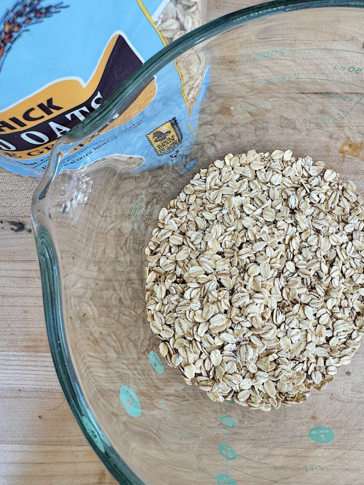 A glass measuring cup filled with oats is placed on a wooden surface next to an opened packet of oats.