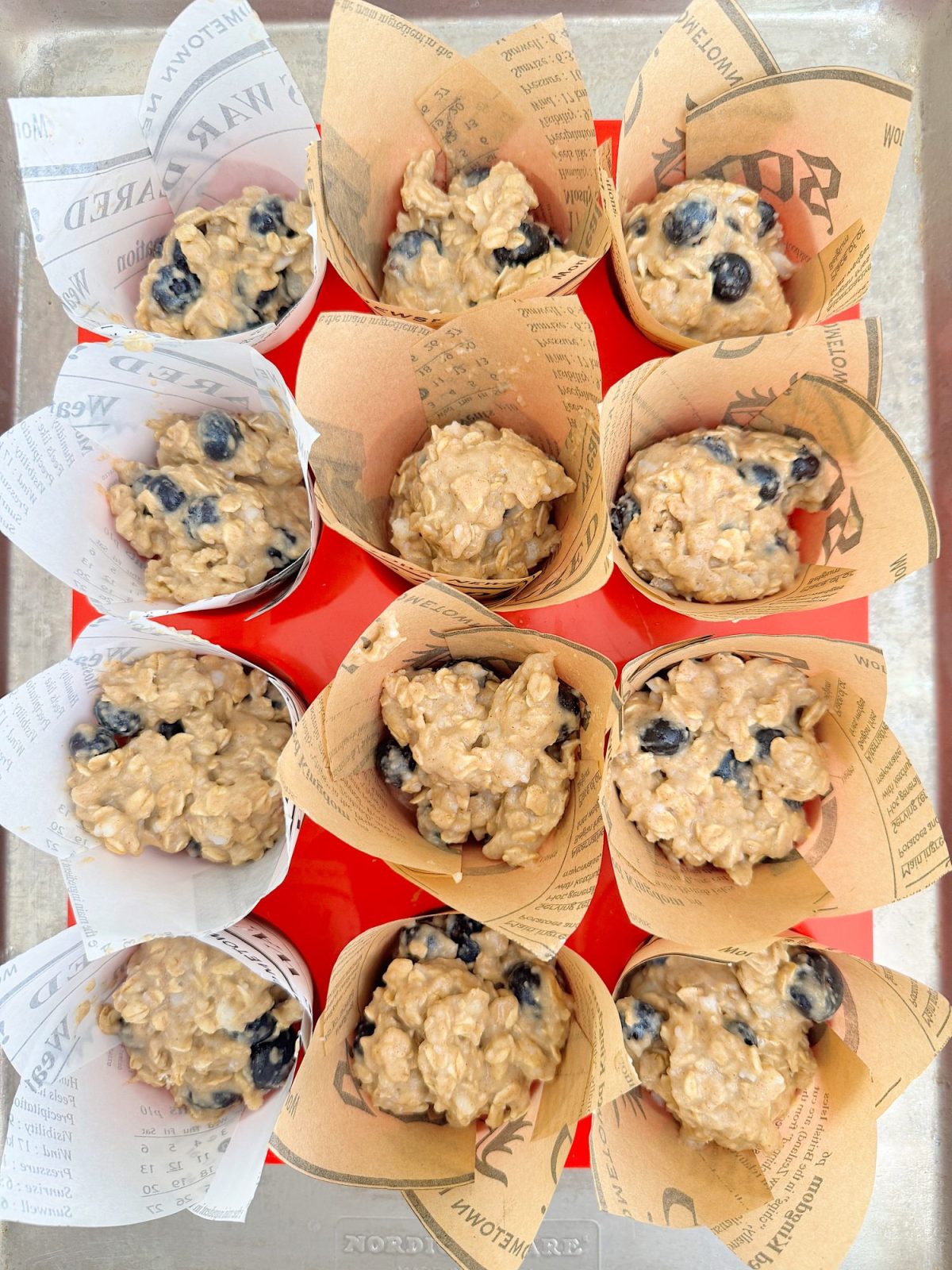 Twelve blueberry oatmeal muffin batters in newspaper-style paper cups, arranged in a metal baking tray before baking.