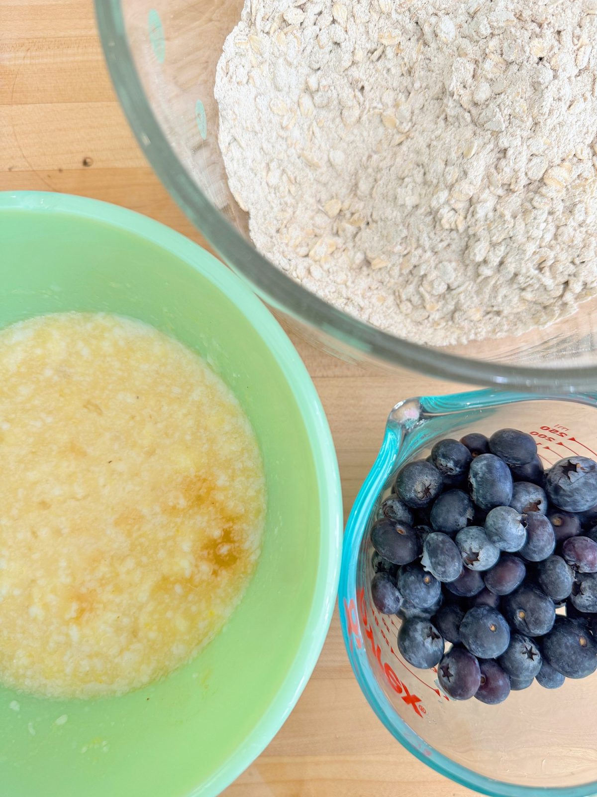 Ingredients for baking on a wooden surface: a bowl with melted butter, a bowl with flour and oats, and a measuring cup filled with blueberries.