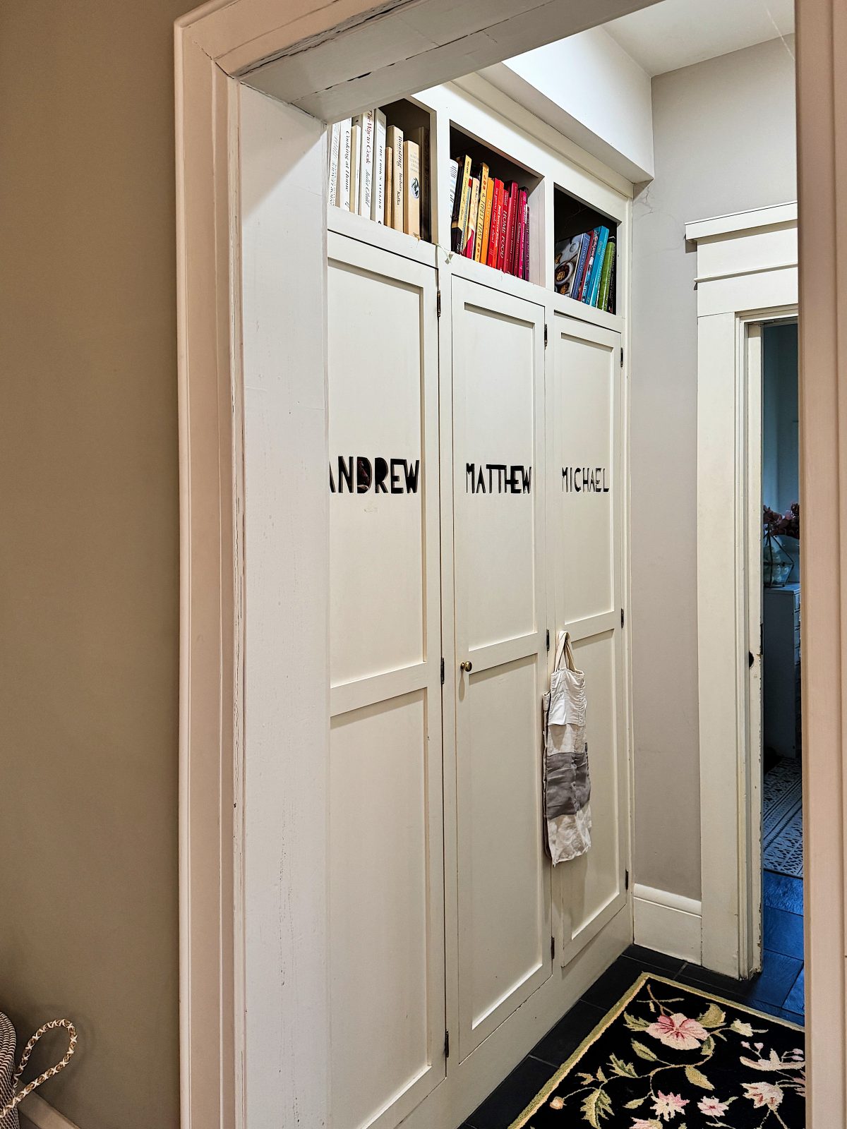 White hallway with three narrow cabinets labeled "ANDREW," "MATTHEW," and "MICHAEL." Some books are visible on top. A patterned rug lies on the floor.