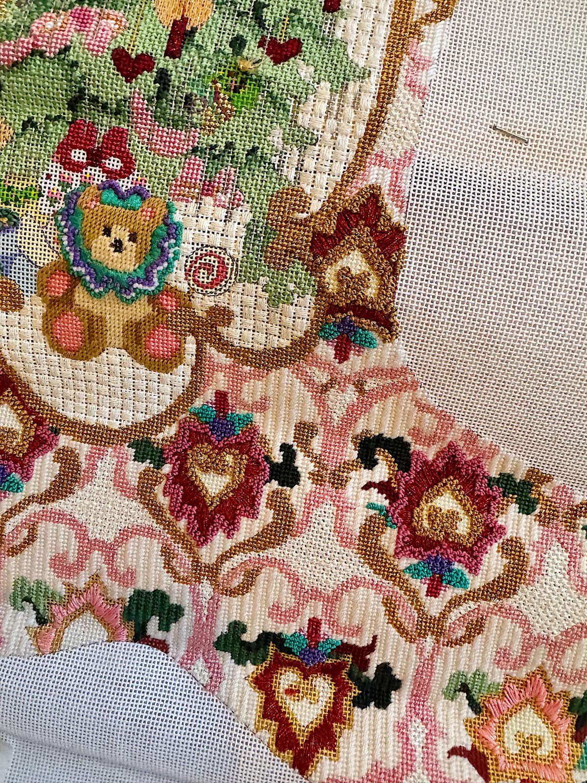 A partially completed cross-stitch project featuring a bear with a tree in the background, surrounded by intricate floral patterns in pink, red, and green tones on a mesh fabric.
