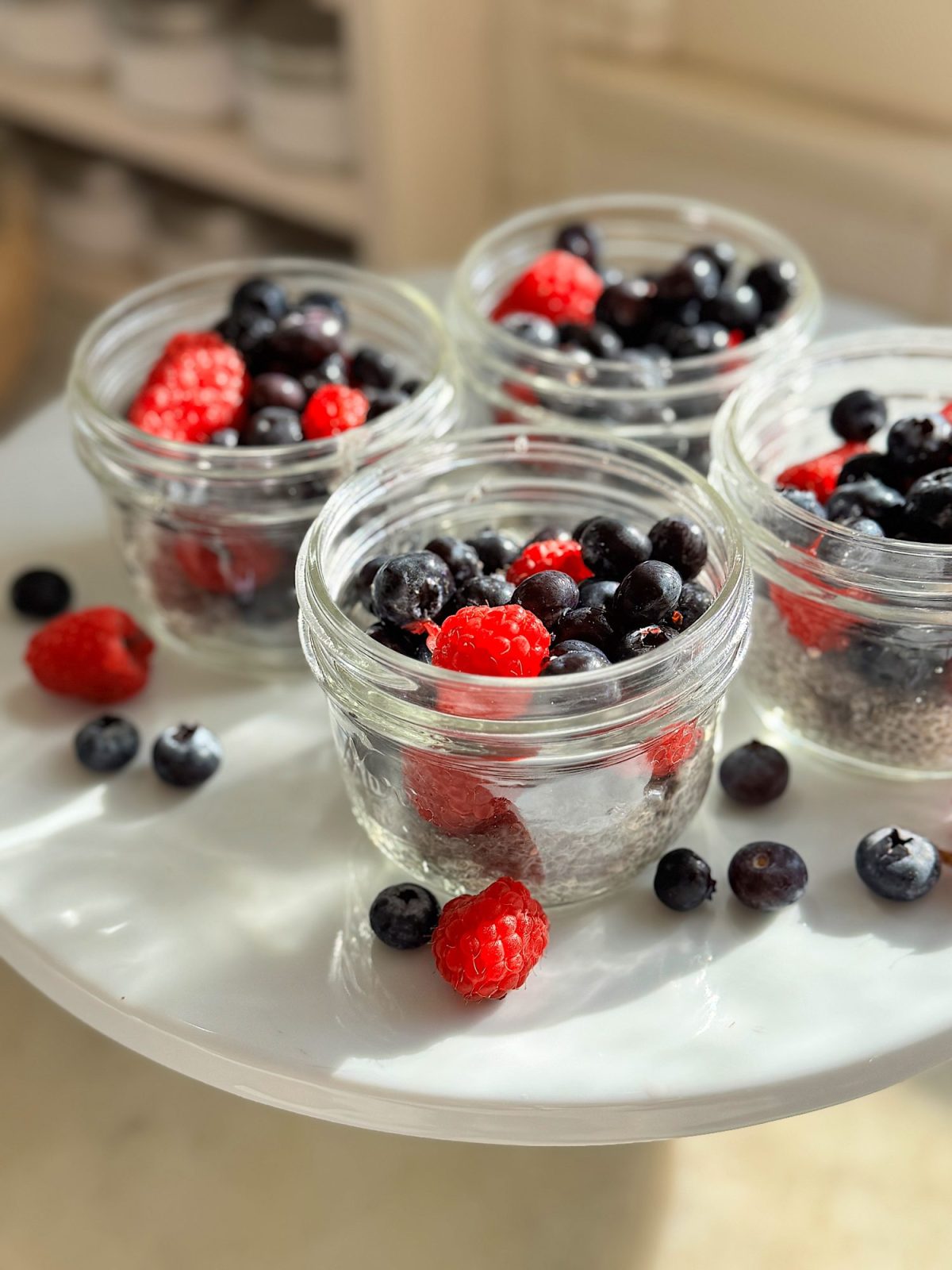 Four glass jars filled with blueberries and raspberries on a white surface, with additional berries scattered around. Sunlight highlights the fruit.