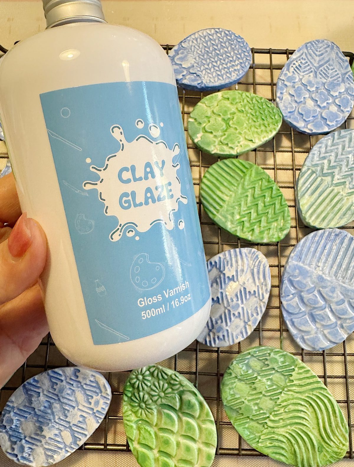 A hand holds a bottle labeled "Clay Glaze" above a wire rack with patterned, painted clay shapes in blue and green.