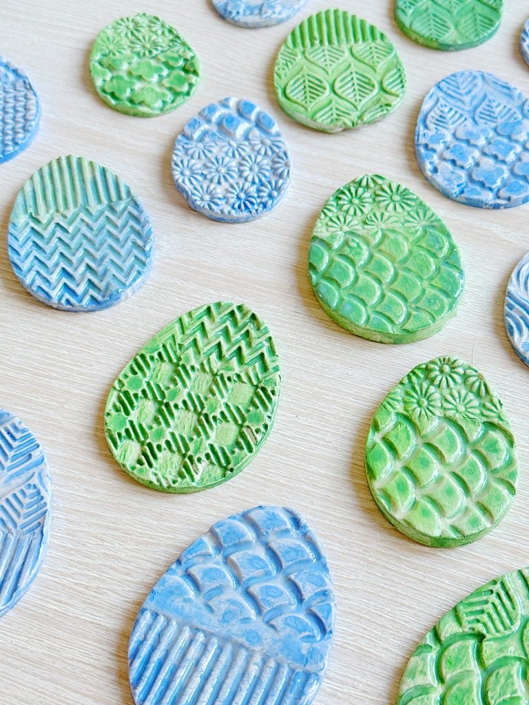 Assorted green and blue oval clay pieces with textured patterns are arranged on a light wood surface.
