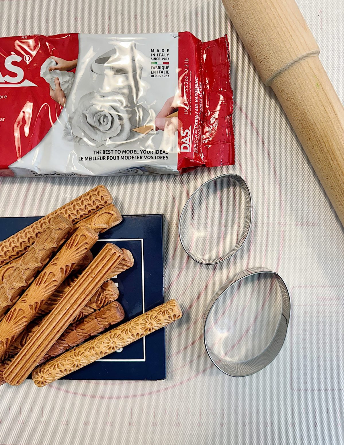 Modeling clay, textured rolling pins, a wooden rolling pin, two oval cutters, and a blue book on a white surface.