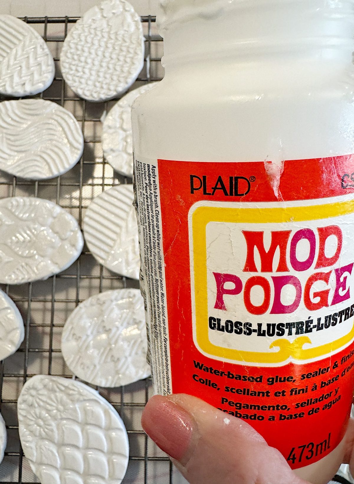 A hand holds a 473ml bottle of Mod Podge gloss sealer. In the background, textured white clay pieces are drying on a wire rack.