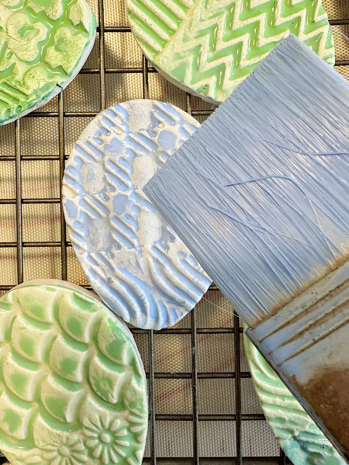 Decorative ceramic pieces with textured patterns in green and white are displayed on a wire rack. A large paintbrush with light blue bristles is shown partially over the ceramics.