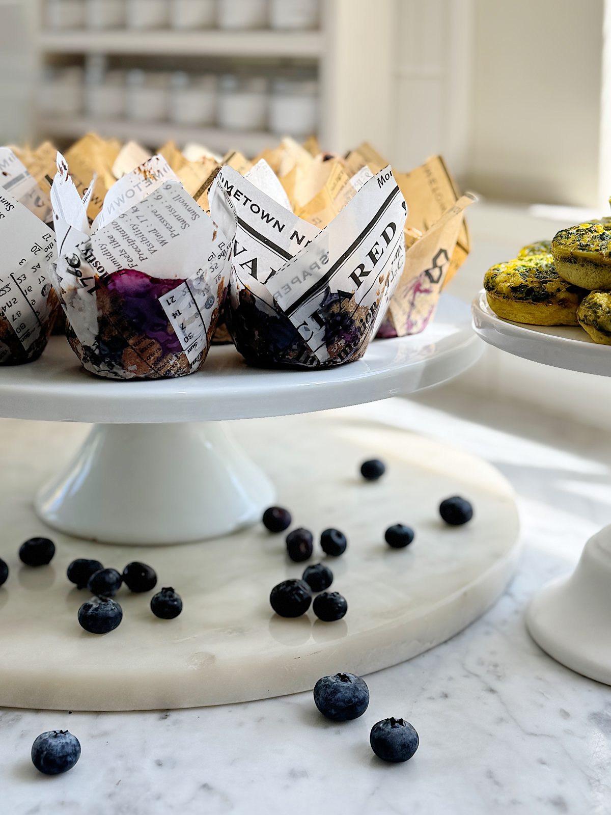 Muffins wrapped in newspaper-style liners and cookies on stands with scattered blueberries on a marble countertop.