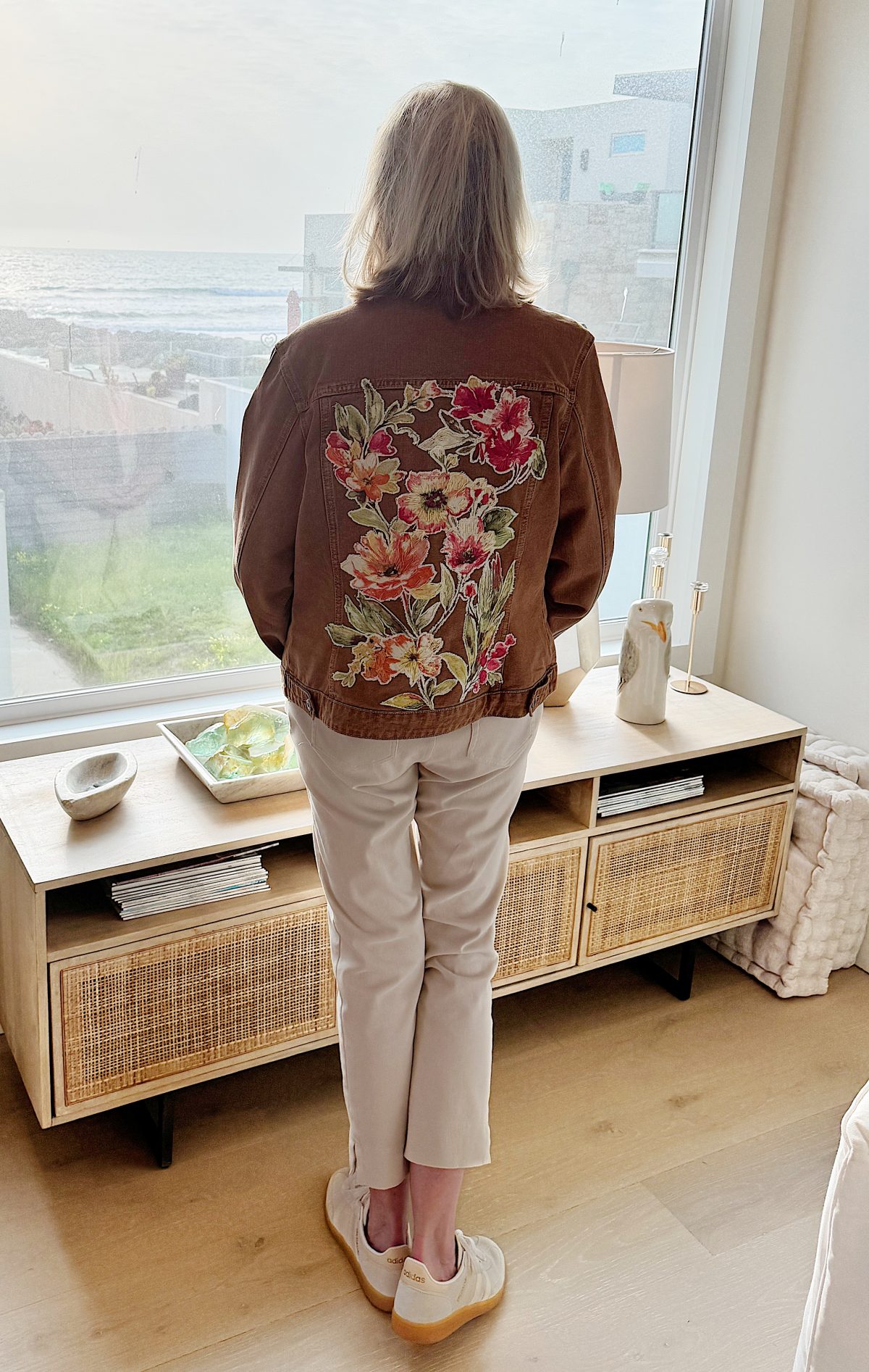 A person stands facing a window, their brown jacket adorned with floral embroidery. With the ambience reminiscent of a filming set, they gaze thoughtfully at the seaside view.