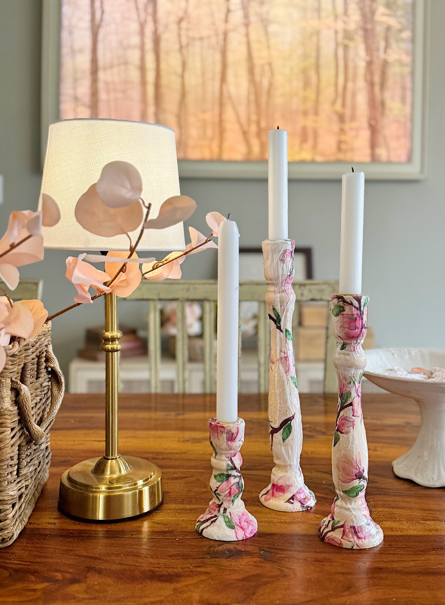 A wooden table holds a brass lamp, a basket with pink leaves, and three floral candle holders with white candles. A blurred forest painting hangs in the background.