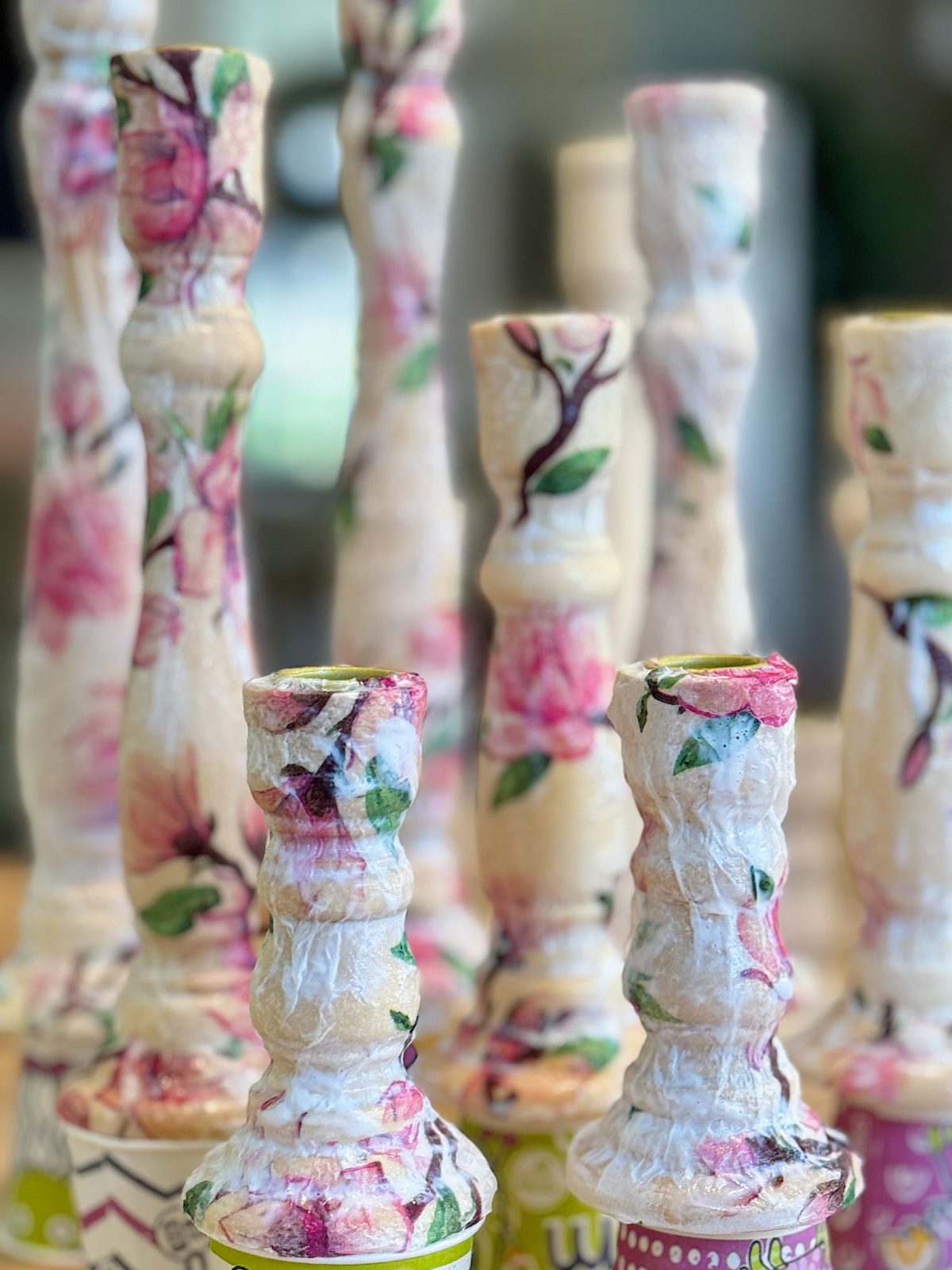 Colorful, handmade candlestick holders with floral patterns arranged in rows, varying in height and adorned with pink and green designs.