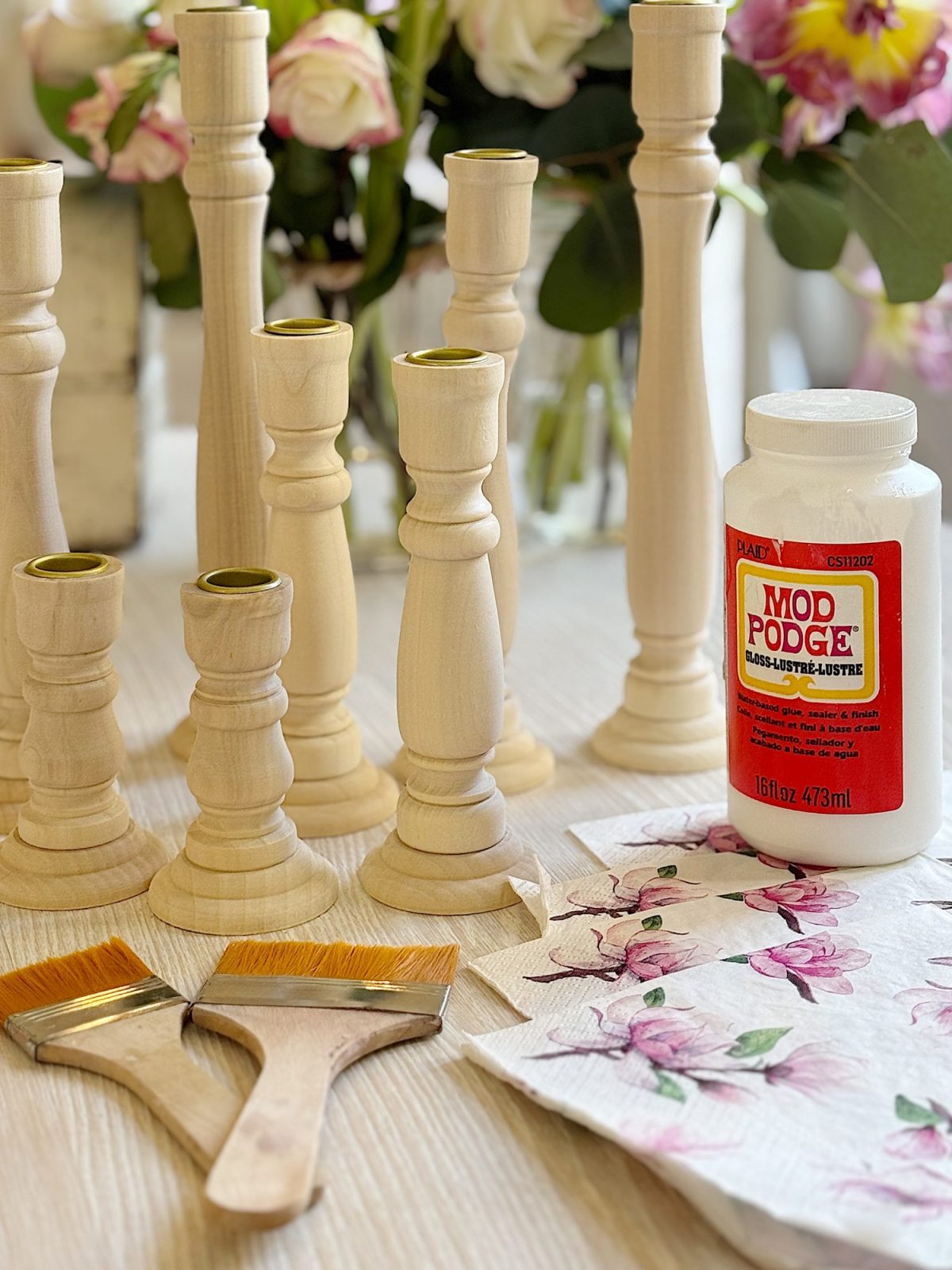 Unfinished wooden candlesticks on a table with two paintbrushes, floral napkins, and a jar of Mod Podge.