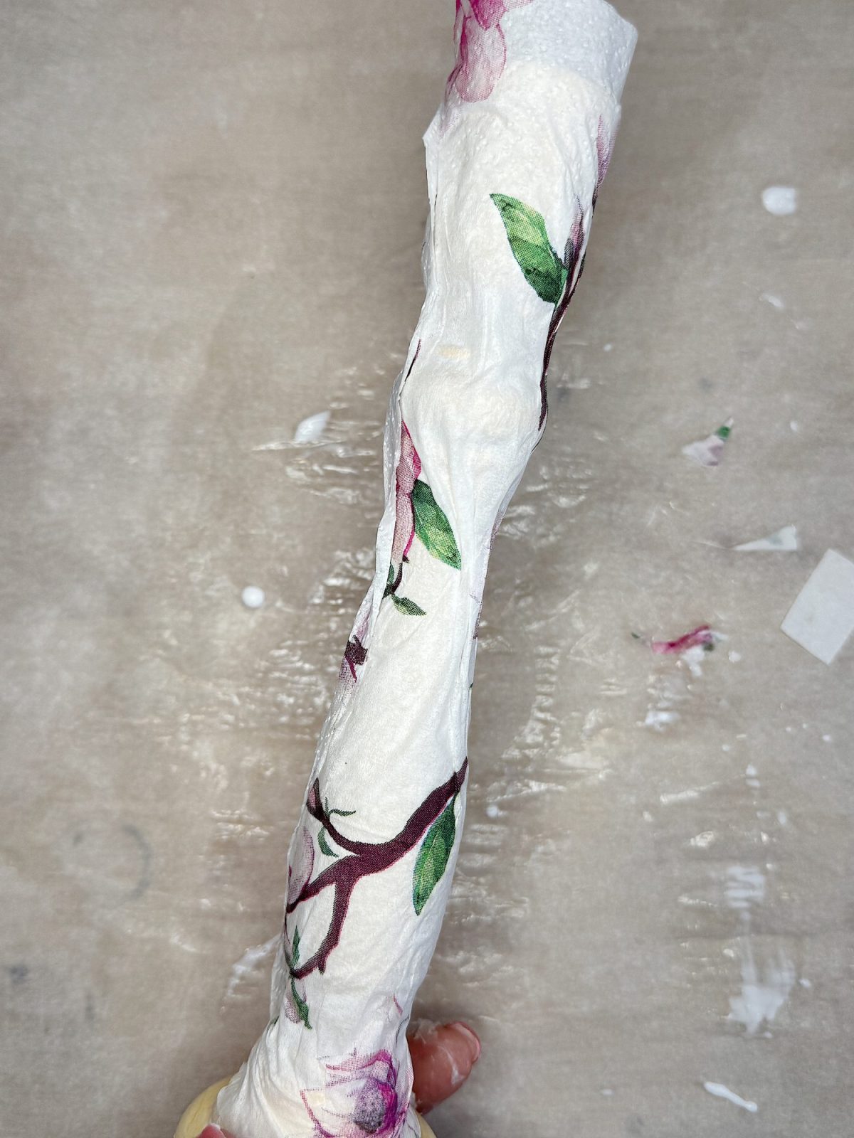 A hand holding a white rolled object decorated with a floral pattern featuring branches, leaves, and flowers, over a neutral background.