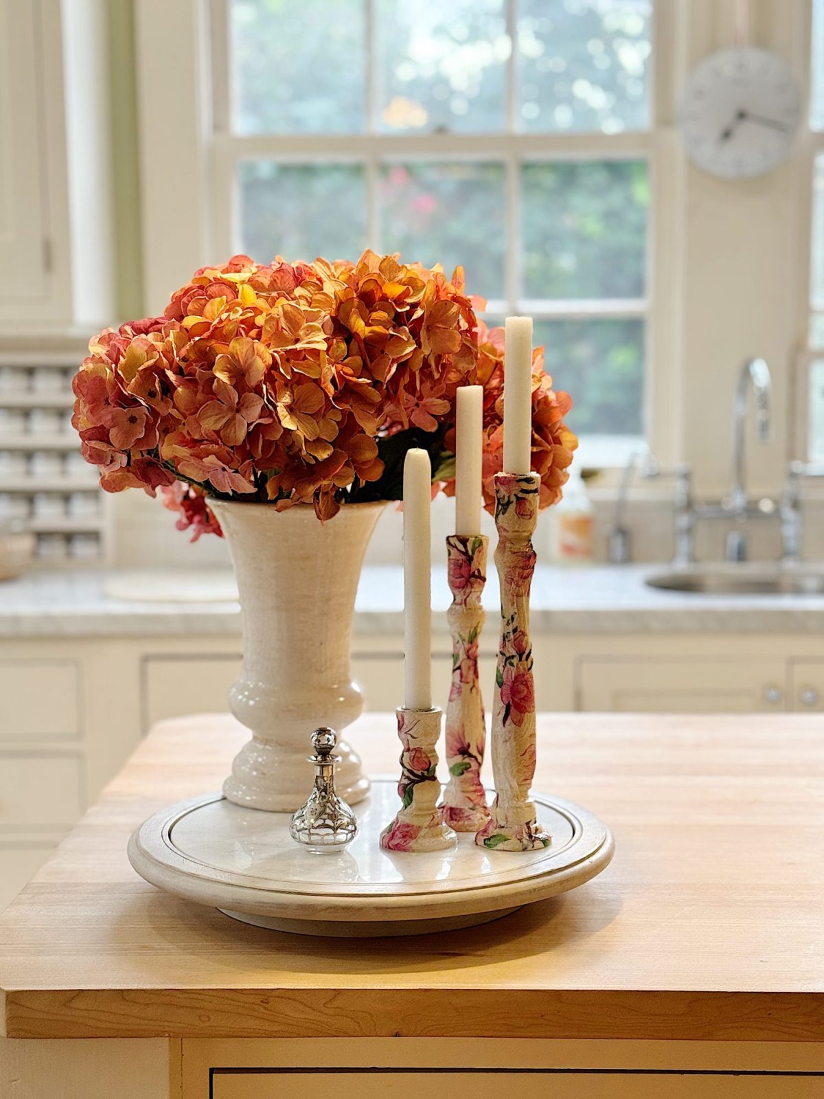 A kitchen island with a vase of orange flowers and three patterned candlesticks. A small decorative item is placed on a round tray beneath a window.