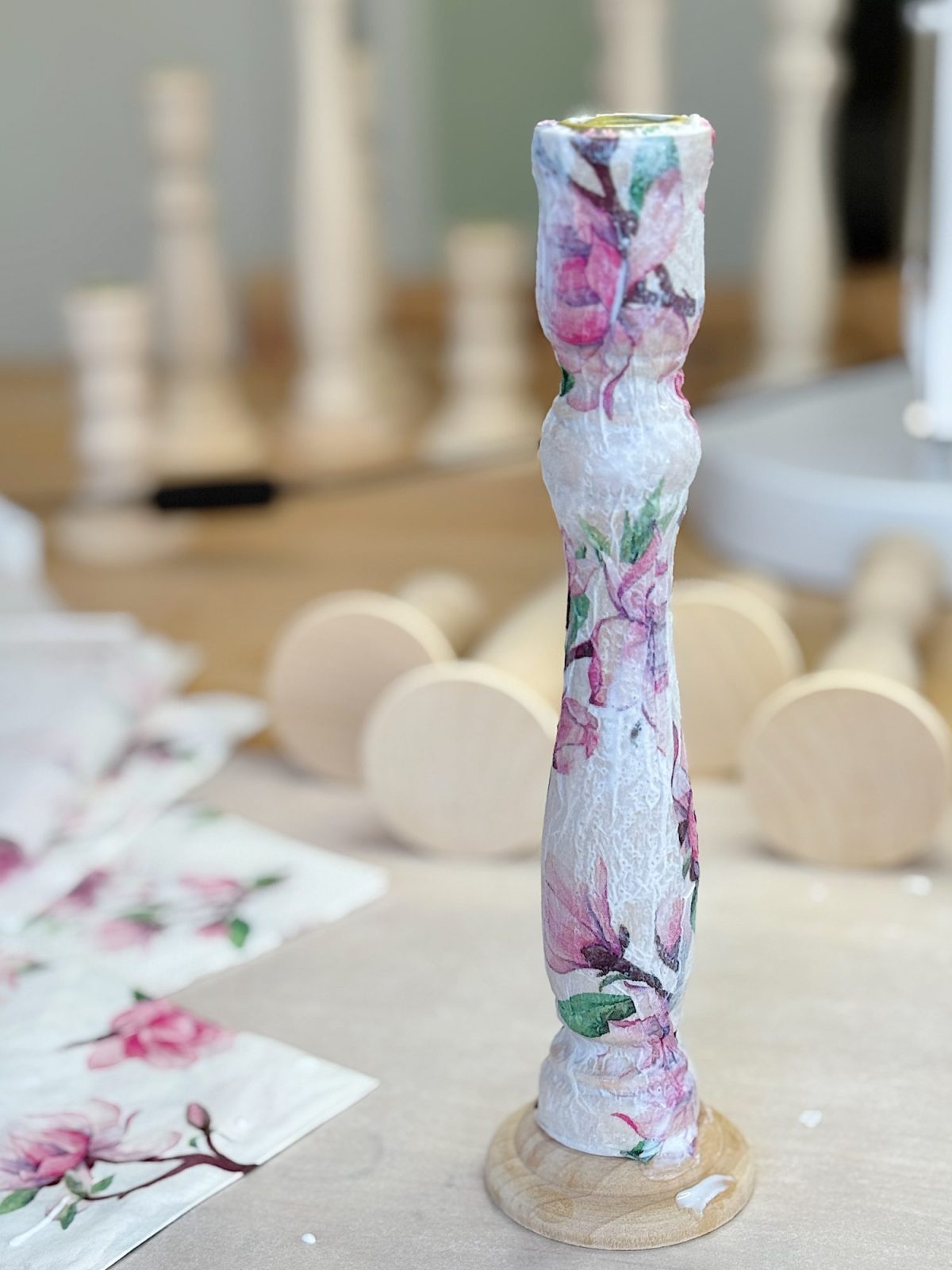 A decorative wooden candlestick holder with a floral design stands on a table, surrounded by crafting materials and additional wooden pieces in the background.