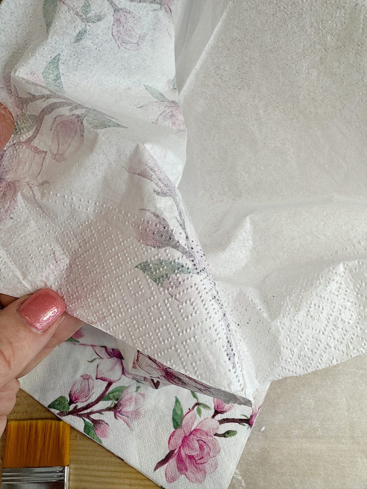 Close-up of a hand holding floral-patterned napkins, revealing multiple layers. The nails are painted pink.