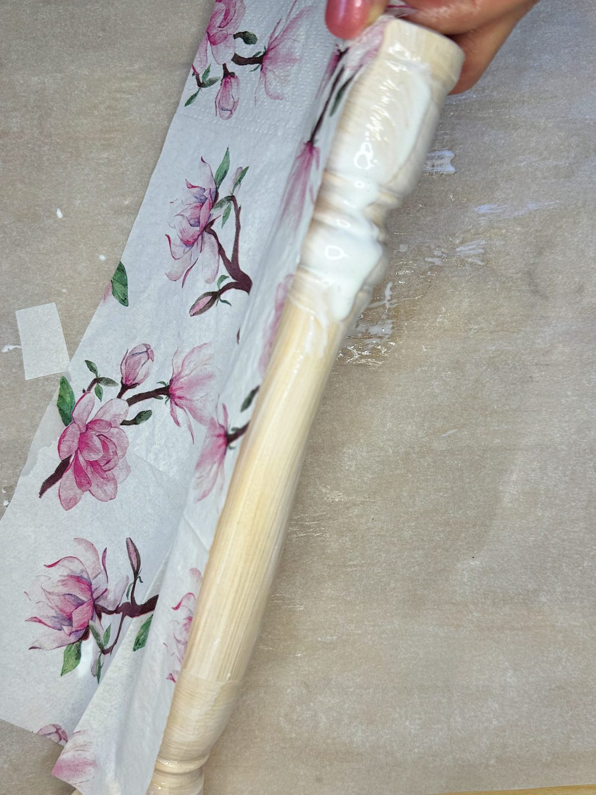 A hand applies a floral-printed fabric onto a wooden spindle using a layer of white glue.