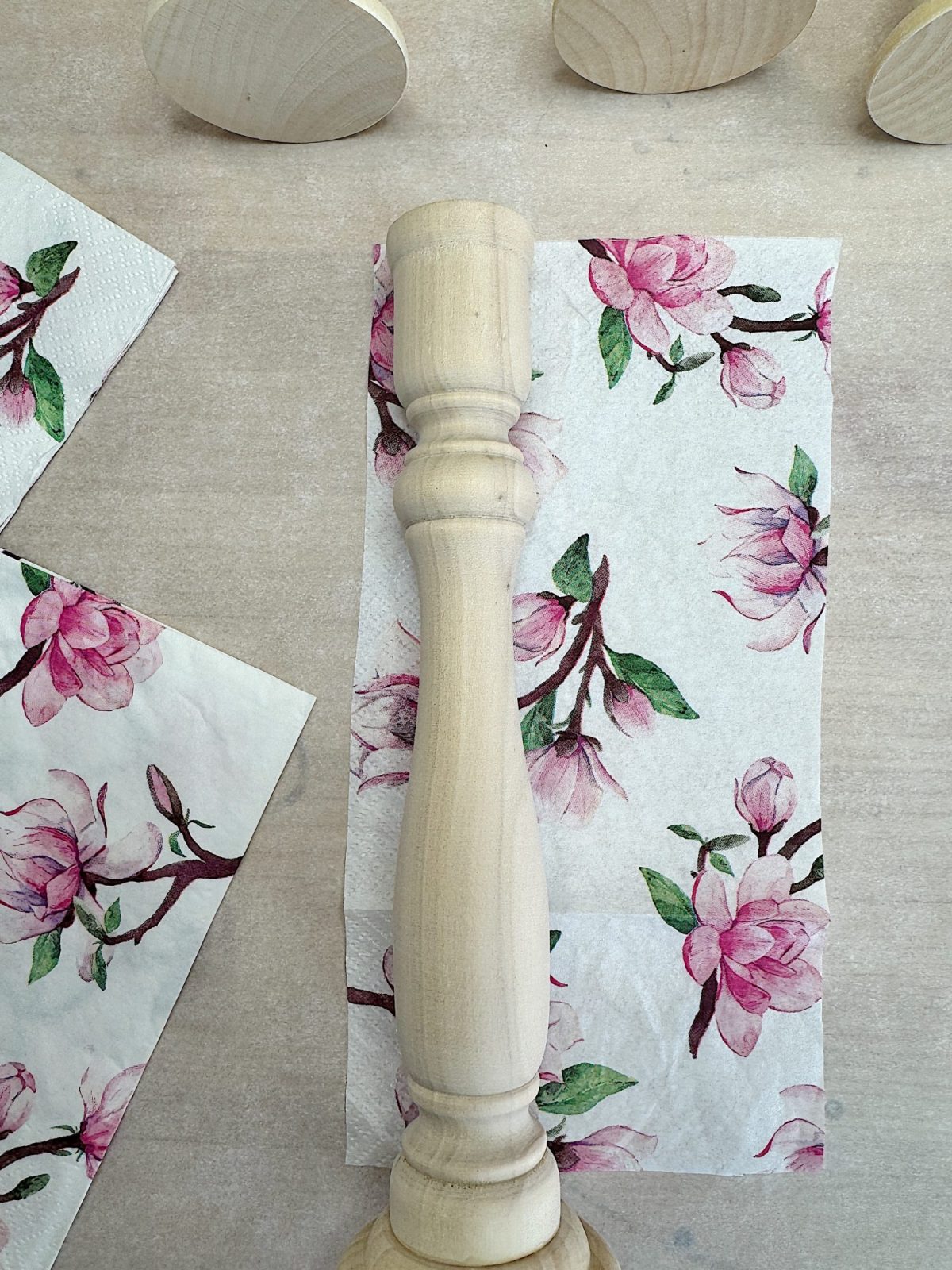 A wooden spindle lies on a napkin with pink floral designs, surrounded by floral-patterned paper pieces on a light wood surface.