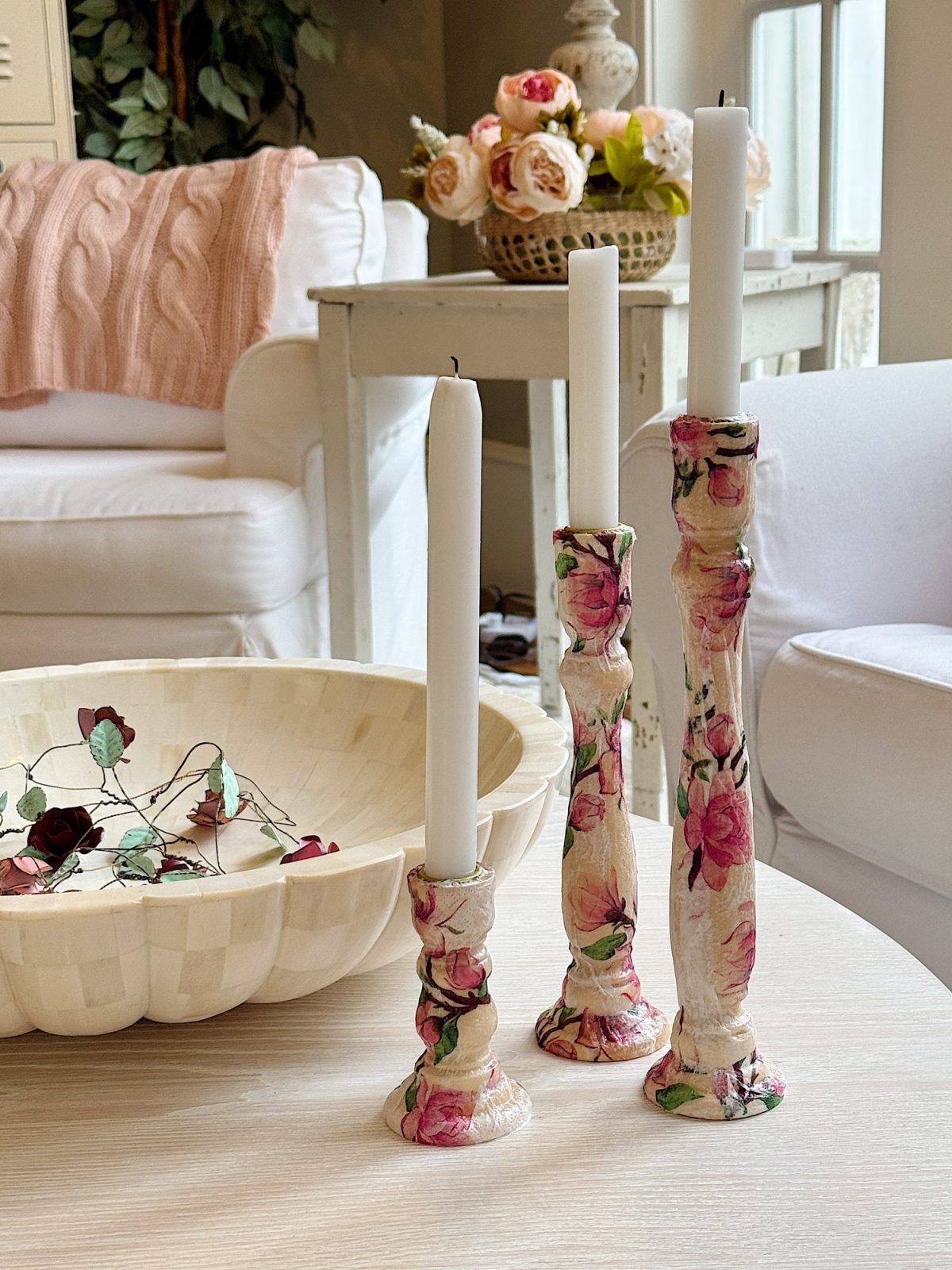 Three floral-patterned candlesticks with white candles on a wooden table, next to a decorative bowl with leaves, in a cozy living room setting with a sofa and a basket of flowers.