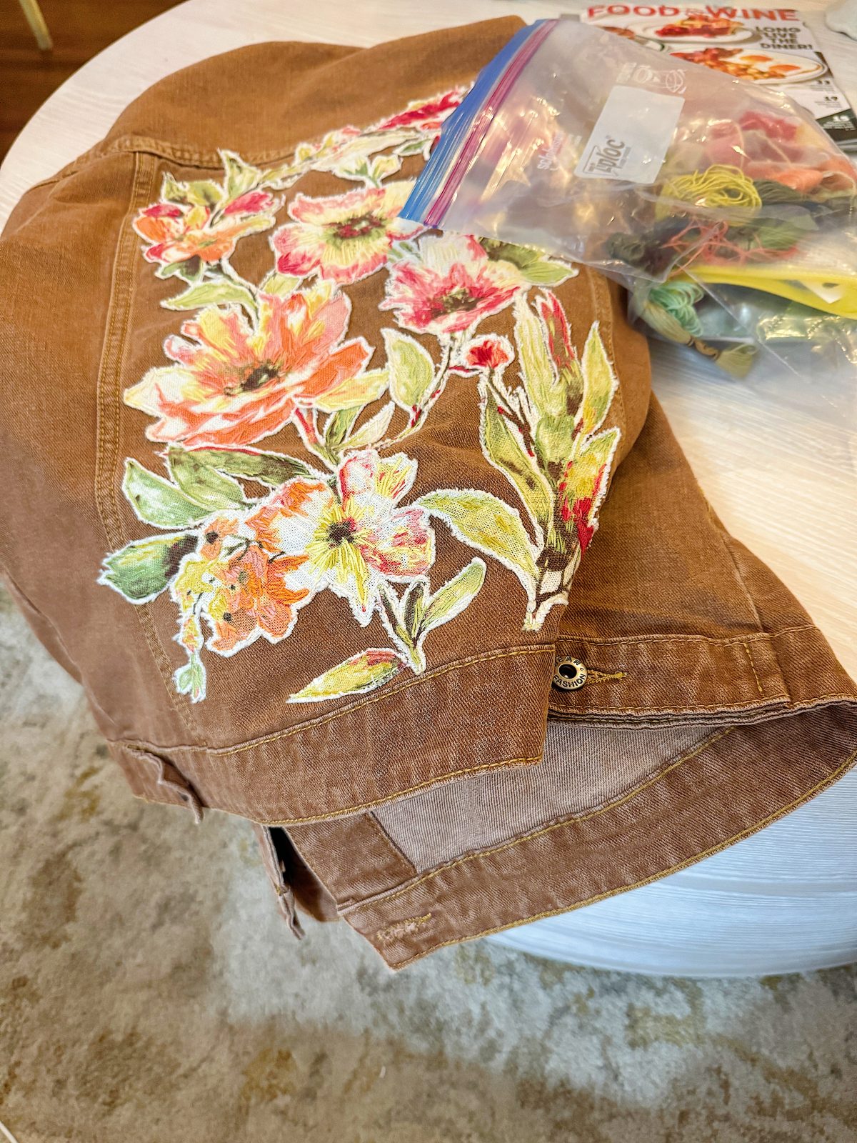 Brown jacket with colorful floral embroidery draped over a table, next to a bag of embroidery thread.