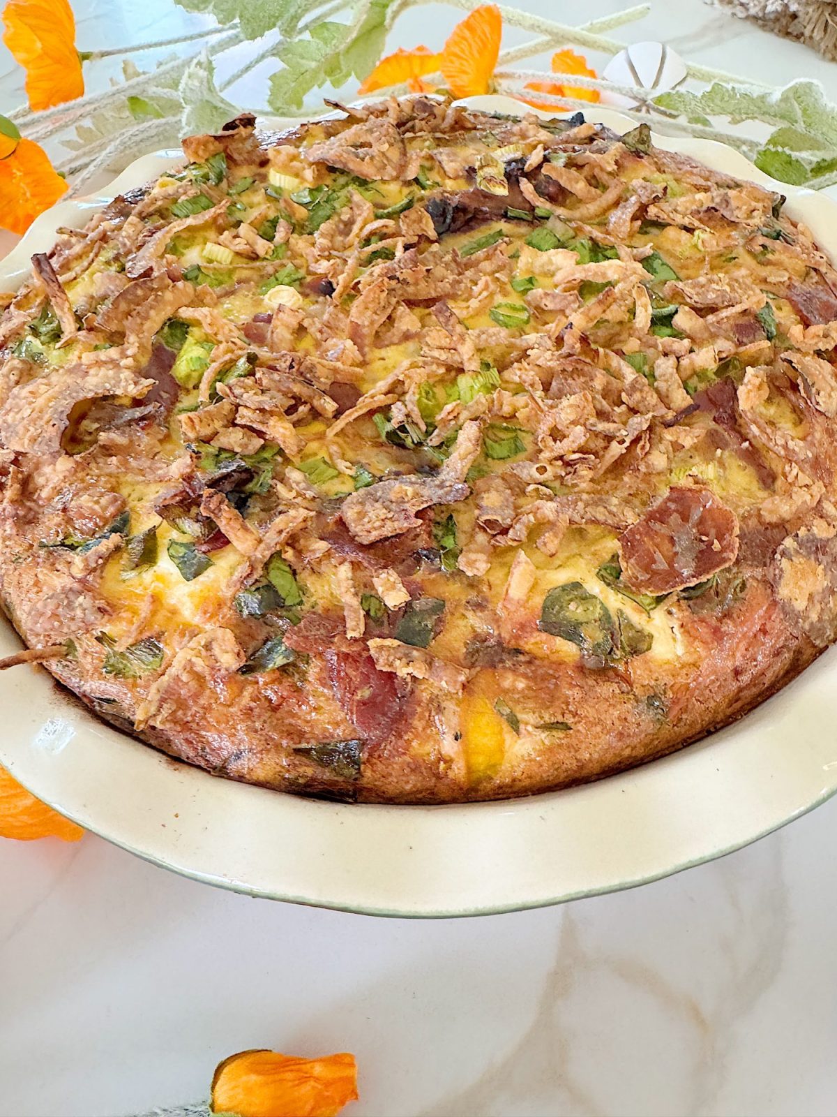 A baked quiche with a golden crust, topped with chopped green onions and crispy fried onions, sits in a ceramic pie dish on a marble surface, surrounded by orange flowers.