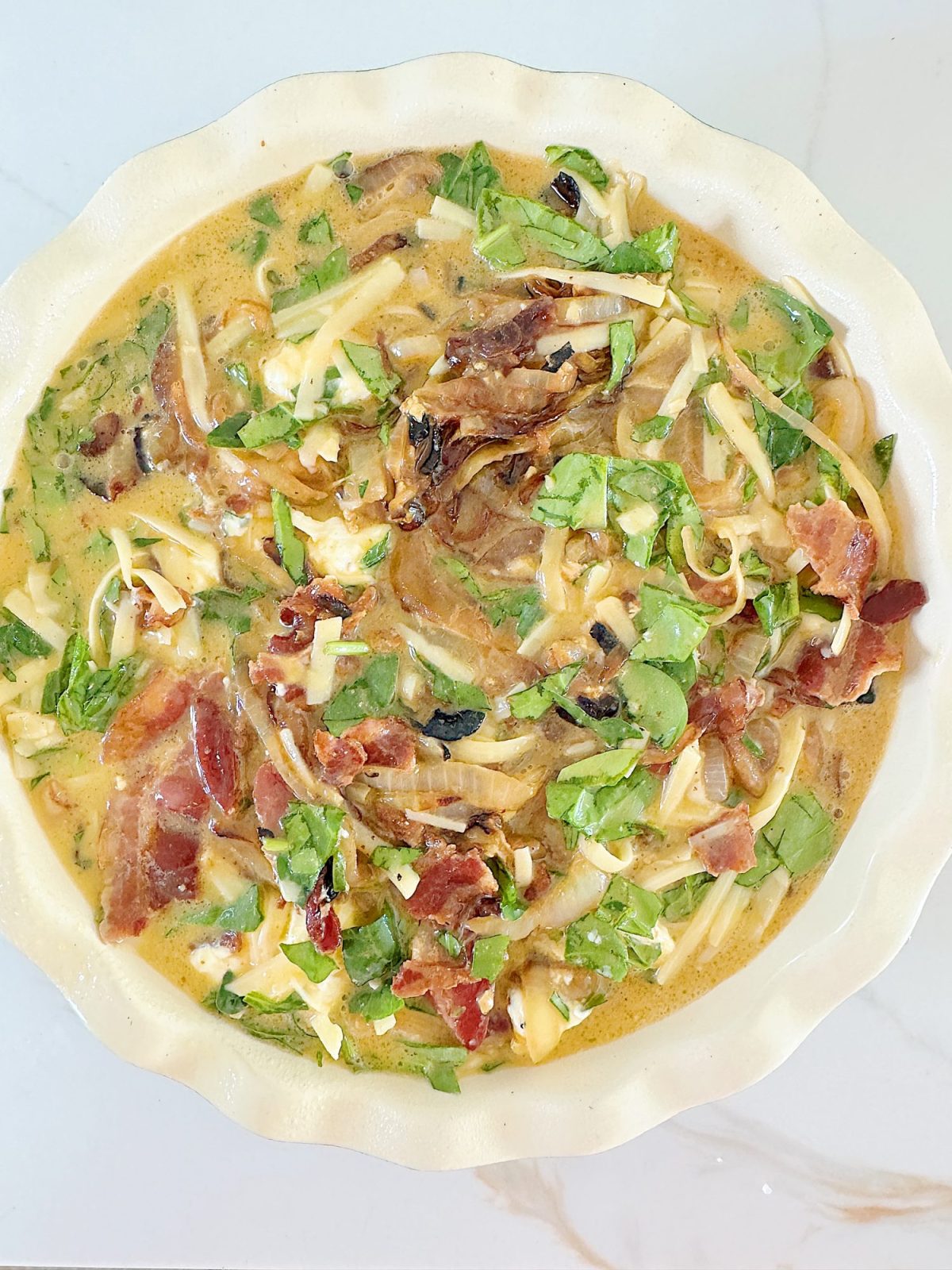 A pie dish filled with creamy soup, garnished with shredded cheese, bacon bits, and chopped greens, on a marble surface.