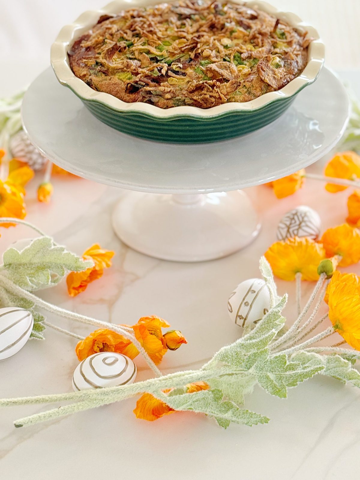 A baked dish on a cake stand, surrounded by orange flowers, greenery, and decorative eggs on a white surface.
