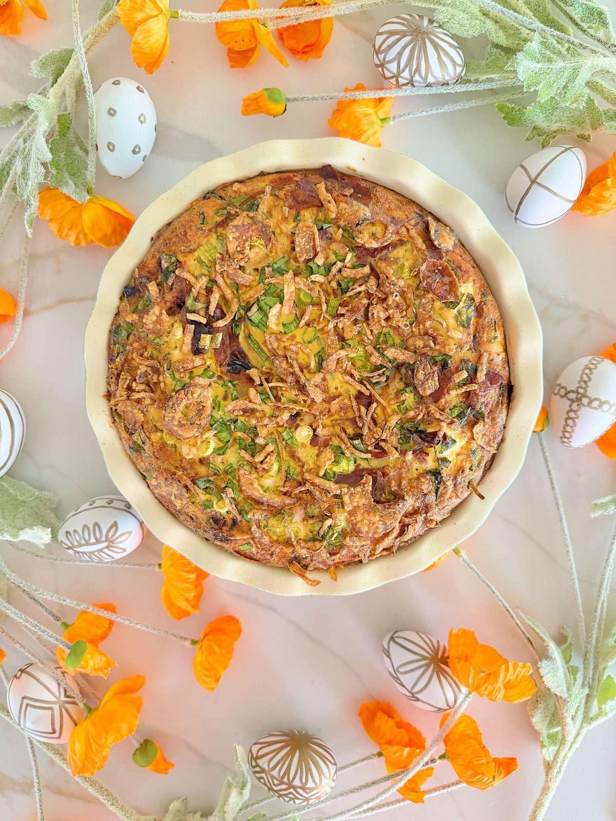 A savory pie in a round dish, placed on a marble surface, surrounded by decorative eggs and orange flowers.