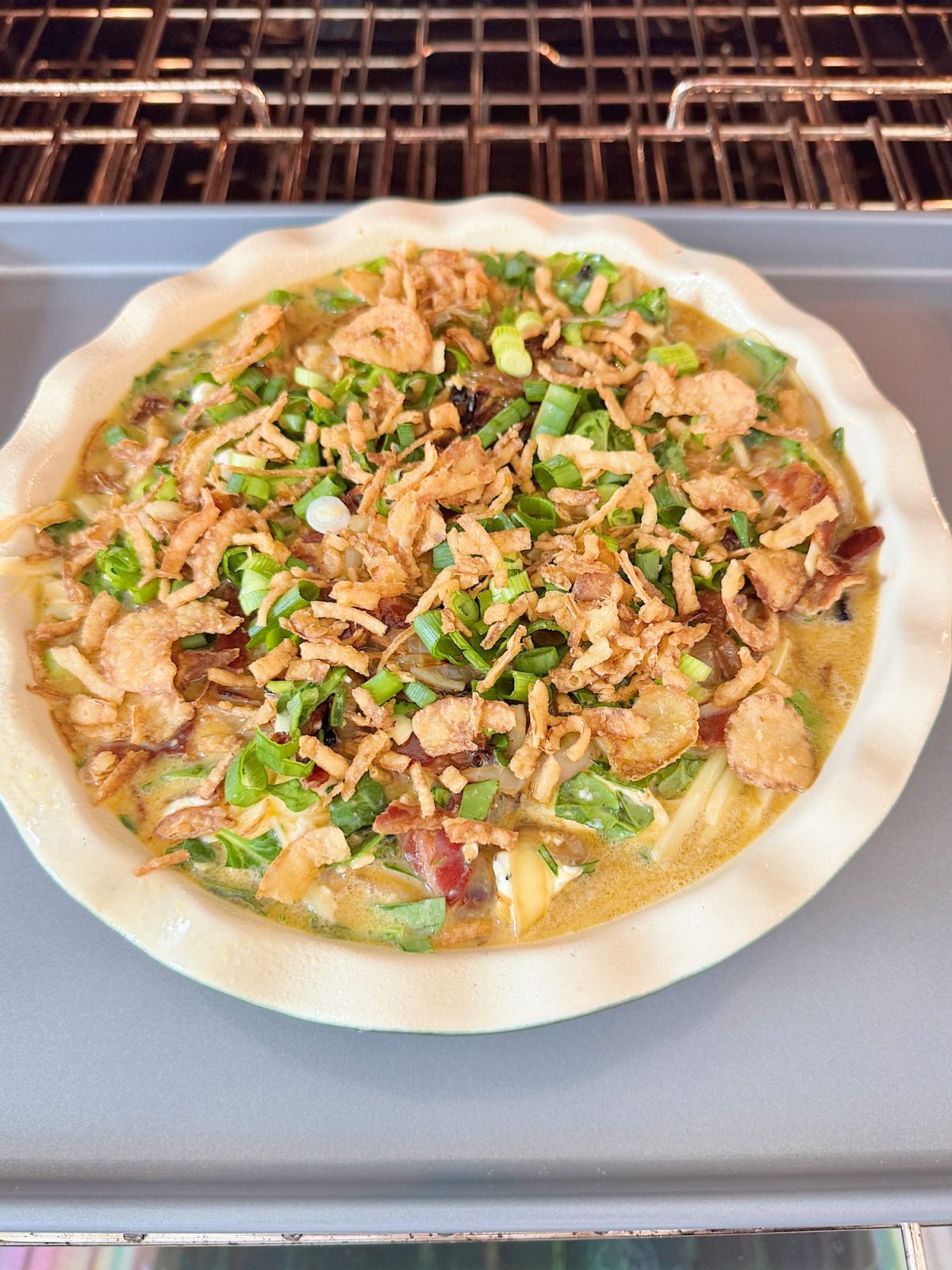 A quiche topped with chopped green onions and crispy fried onions is baking in the oven on a baking sheet.